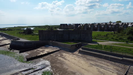 A-rising-drone-shot-of-an-abandoned-military-garrison-situated-behind-a-seawall-on-the-edge-of-beach-and-mudflats-on-a-tidal-estuary-at-low-tide,-on-a-clear-and-hot-summer-day