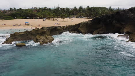 Olas-Del-Mar-Rompiendo-En-La-Costa-Rocosa-Con-Gente-Nadando-En-La-Playa-La-Poza-Del-Obispo-En-Arecibo,-Puerto-Rico