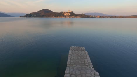 Peaceful-establishing-shot-of-concrete-jetty-on-lake-Maggiore-calm-water-in-Italy,-Angera-castle-in-background