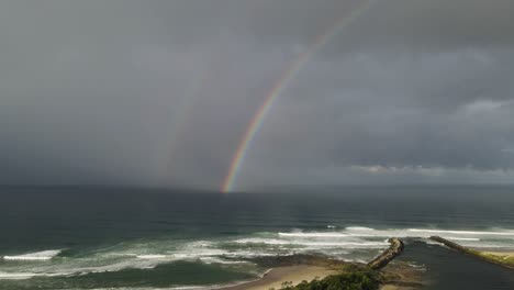 River-mouth-leading-out-to-a-popular-surfing-beach-with-a-dazzling-rainbow-and-rain-clouds-above-a-turbulent-ocean-swell