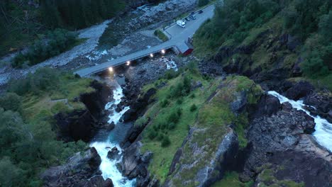 Top-of-Latefoss-waterfall-looking-down-at-road-and-bridge-with-lights-during-dusk-hour---Downward-moving-aerial-in-middle-of-waterfall