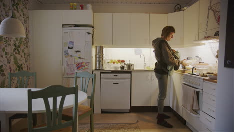Mother-carrying-her-baby-on-a-sling-moves-around-kitchen-cooking,-wide-shot