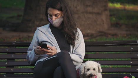 Woman-wearing-a-mask,-sitting-by-her-dog-on-park-bench-using-her-phone