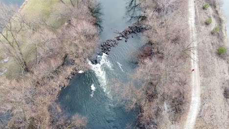 Small-river-dam-and-rapids-on-scenic,-idyllic,-willow-tree-lined-river-in-winter-with-a-person-wearing-red-running-on-the-track-in-rural-countryside-of-Boise,-Idaho,-USA
