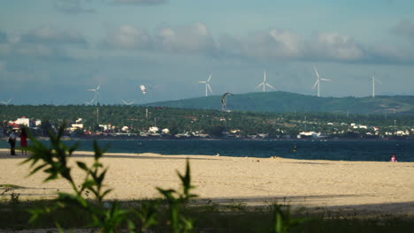 A-seaside-view-of-developed-suburbs-with-wind-energy-farm-running-in-the-background-and-visitors-at-the-beach-enjoying-kitesurfing,-doing-photography