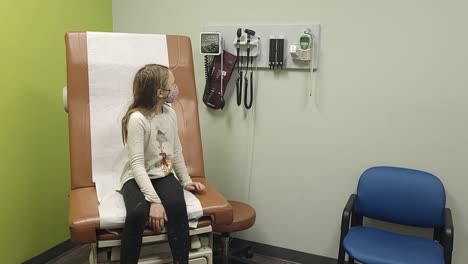 Junges-Mädchen-Sitzt-In-Der-Arztpraxis-Und-Schaut-Sich-Neugierig-Um,-Während-Es-Wartet-Und-Eine-Gesichtsmaske-Trägt