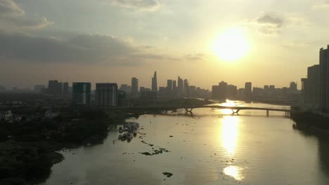 tracking-across-the-Saigon-river,-Vietnam-from-drone-with-view-to-bridge-and-modern-city-skyline-at-sunset