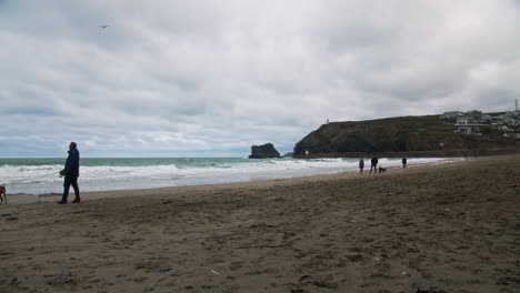 People-Walk-on-Portreath-Beach-in-Winter-with-Pet-Dogs-as-Waves-Crash-on-Coastal-Shore-of-Cornwall
