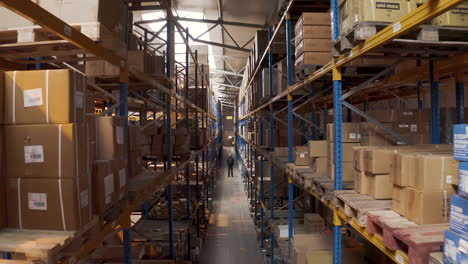 Man-standing-in-a-long-storage-warehouse-aisle-with-boxes,aerial-zoom