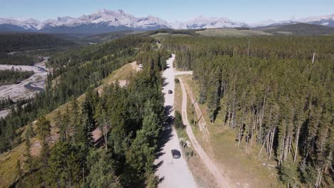 Multiple-cars-driving-along-a-dusty-logging-road-through-a-boreal-forest-with-a-majestic-Rocky-Mountain-backdrop