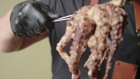 A-male-chef-wearing-black-gloves,-hold-up-a-half-cooked-octopus-with-a-tong-from-a-chopping-board,-slow-motion-shot