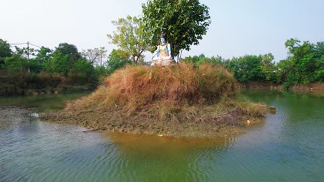 Aerial-drone-rotated-view-of-the-small-grassy-island-where-the-blue-statue-of-Hindu-God-Shiva-rests,-with-a-background-view-of-a-white-vacation-house-and-a-large-lake-in-Vadodara,-India