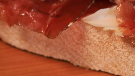 Close-up-view-of-oily-high-quality-spanish-serrano-ham-with-bread-rotating-on-wooden-board,-macro-shot-from-above