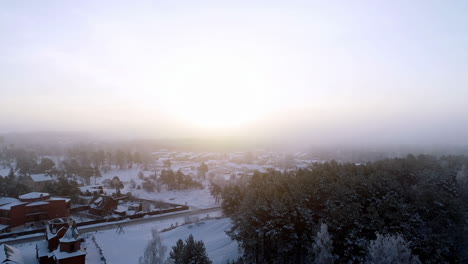 Aerial-view-over-the-beautiful-small-town-houses-beside-the-forest-covered-with-snow-on-a-foggy-winter-morning