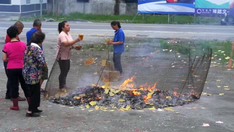 Asian-men-and-women-throwing-paper-money-into-caged-fire-pit-during-spiritual-ceremony-to-appease-ancestors-and-ghosts-filmed-in-slow-motion