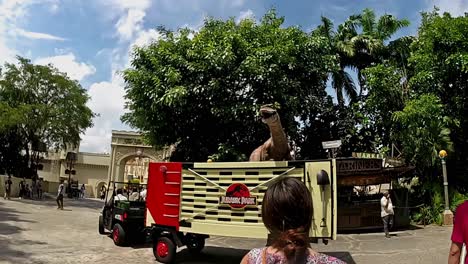Touristen-Beobachten-Amüsiert,-Wie-Eine-Animierte-Dinosaurier-Nachbildung-In-Einem-Lastwagen-In-Einem-Teil-Der-Universal-Studios-Singapur-Vorbeifährt