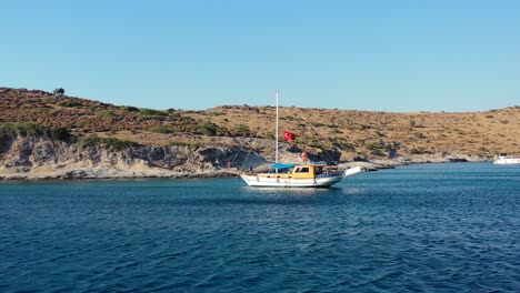 Luftdrohne-Umkreist-Ein-Türkisches-Segelboot-Mit-Touristen,-Das-In-Der-Blauen-Bucht-Des-Ägäischen-Meeres-In-Der-Nähe-Von-Bodrum,-Türkei,-Vor-Anker-Liegt,-Während-Die-Sonne-über-Den-Hügeln-Untergeht
