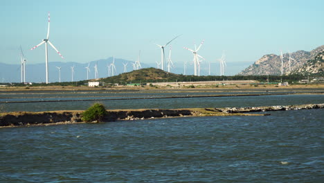 Turbinas-Eólicas-Girando-En-Un-Parque-Eólico,-Agua-Del-Lago-Del-Río-Fluyendo-En-Primer-Plano