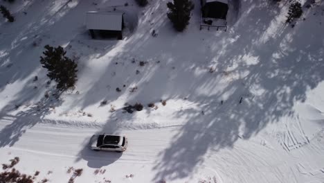 Family-leaving-mountain-cabin-on-snow-covered-backcountry-road-during-the-day,-top-down-aerial