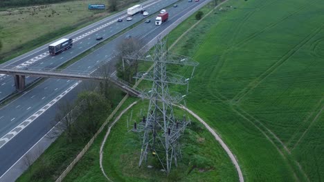Vehicles-on-M62-motorway-passing-pylon-tower-on-countryside-farmland-fields-aerial-high-view-orbit-right