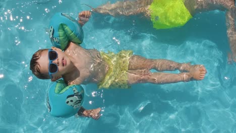 Entspannter-Kleiner-Junge-Mit-Sonnenbrille,-Der-Mit-Bunten-Schwimmkörpern-Im-Schwimmbad-Schwimmt