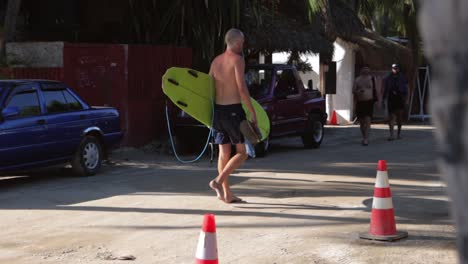 Caucasian-shirtless-male-surfer-with-a-green-surf-board-walking-barefoot-on-muddy-dirt-road-in-Mexico-after-catching-waves