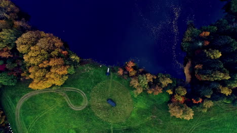 Aerial-top-shot-over-rectangular-cabin-beside-a-pristine-lake-surrounded-by-autumnal-trees-in-rural-landscape-at-daytime