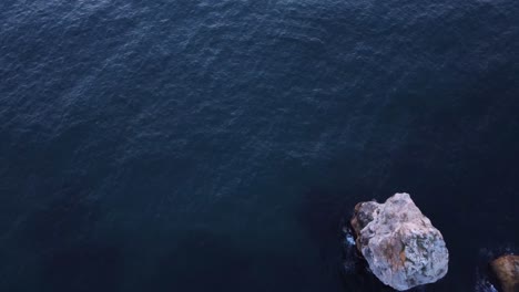 Top-down-tilt-aerial-view-of-waves-splash-against-rocky-seashore,-background