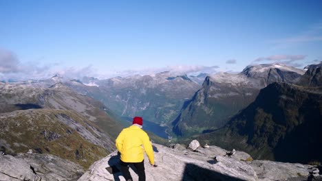 Male-hiker-with-a-yellow-jacket-and-red-hat-walking-to-the-edge-of-mountain-cliff-to-enjoy-a-stunning-view