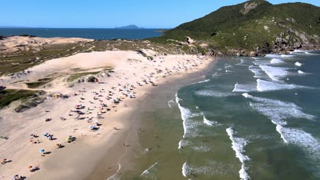 Aerial-drone-scene-of-urbanized-paradise-beach-with-people-walking-on-the-sand-with-dunes-mountains-and-sea-beach-of-santinho-FlorianÃ³polis-in-the-late-afternoon