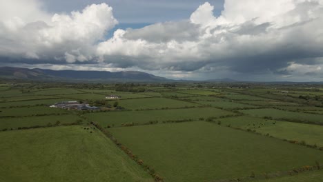 Drone-shot-of-a-vast-rural-Irish-landscape-with-green-fields,-a-farm-and-hills-in-the-distance