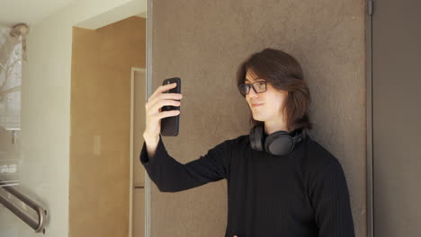 Young-man-with-glasses-on-a-video-call-using-his-mobile-phone,-headphones-around-his-neck