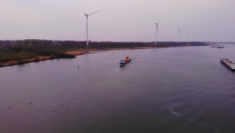Aerial-View-Of-Martie-Cargo-Ship-Approaching-On-Oude-Maas-With-Birds-Resting-On-Rivers-Surface-And-Still-Wind-Turbines-In-Background