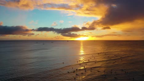 Colorful-Sun-Setting-Over-Group-of-Surfers-and-Boats-Sailing-At-Dawn-On-Waikiki-Beach-in-Honolulu-Hawaii-With-Calm-Ocean-Waves---aerial-drone-shot