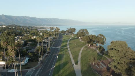 High-altitude-aerial-over-coastline-city-park-during-sunset-with-trees-and-the-Pacific-ocean-with-mountains-in-the-background,-in-Santa-Barbara,-California,-USA
