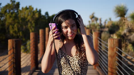 Eine-Attraktive-Junge-Hispanische-Frau-Geht-Und-Hört-Musik-über-Ihre-Kopfhörer-Mit-Einem-Smartphone,-Während-Sie-In-Zeitlupe-Lacht-Und-Lächelt