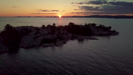 Reveal-of-Epic-Sunset-over-Rocky-Pine-Tree-Island-in-Blue-Lake,-Drone-Aerial-Wide-Dolly-Out