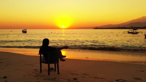 Silhouette-of-a-woman-relaxing-in-the-chair-on-the-sandy-beach-during-the-golden-sunset
