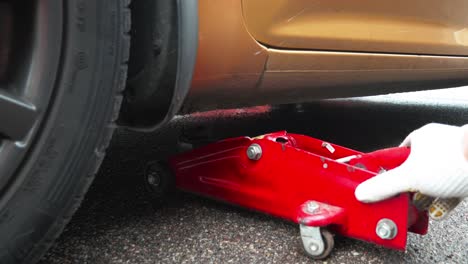 Man-hands-pushing-a-red-car-jack-under-gold-color-car-for-wheel-replacement