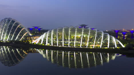 Singapore-night-to-early-morning-drone-hyper-lapse-moving-left-to-right-over-the-water-featuring-Gardens-by-the-bay,-Marina-Bay-Sands,-Super-trees,-Cloud-Dome,-Flower-Dome-with-mirror-reflection