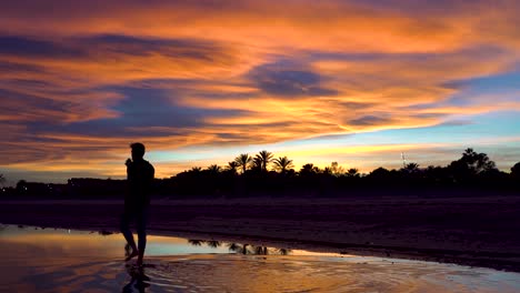 Man-walking-on-the-water-towards-the-bottom-of-the-beach,-the-cloudy-sky-with-beautiful-colors-at-sunset-on-the-background-of-the-image