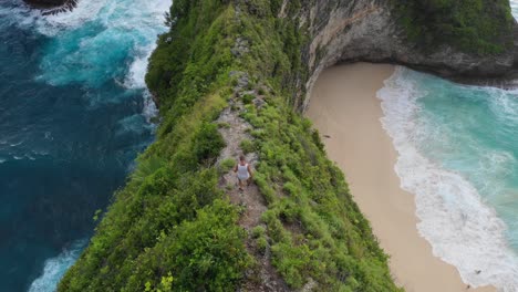 Man-hiking-down-sharp-cliff-with-idyllic-beach-in-background,-Aerial,-Bali