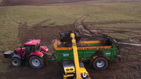 AERIAL:-Reveal-Shot-of-Yellow-Tractor-Loading-Steaming-Hot-Manure-into-Trailer-of-Another-Heavy-Machinery