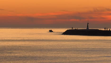 Motor-boat-at-dawn-on-calm-sea-passing-harbor-wall-with-people-fishing