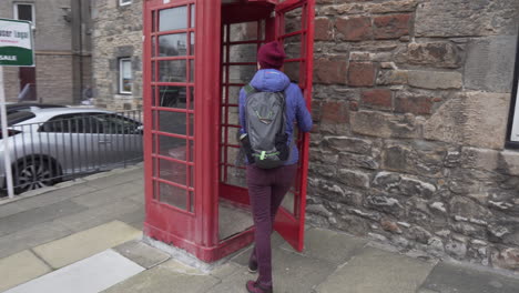 Dolly-circling-shot-of-girl-walking-to-and-entering-a-UK-standard-telephone-booth-in-city-of-Edinburgh,-Scotland