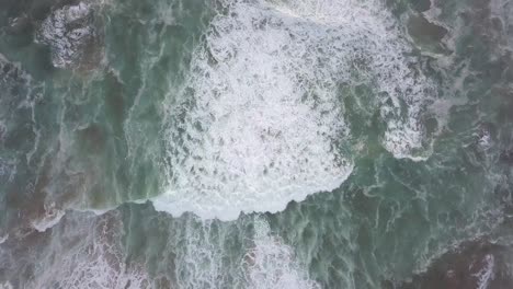 Churning-white-foamy-waves-move-slowly-shoreward,-vertical-aerial
