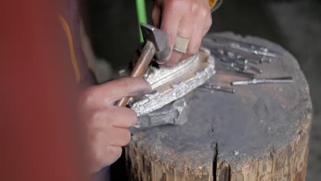 A-skilled-craftswoman-carefully-carving-design-with-a-hammer-and-chisel-on-a-small-silver-mound-on-a-small-log---Close-up