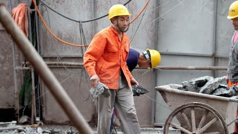 A-construction-worker-carries-debris-as-they-demolish-concrete-floor-at-a-site-using-demolition-hammer-and-a-wheelbarrow,-Slow-motion-handheld-shot-worker-looks-in-camera-direction