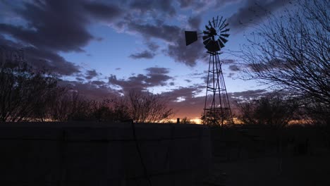 Timelapse-of-a-windpump-and-water-tank-against-a-dramatic-sunset-with-orange-clouds-transitioning-into-stars-on-a-sheep-farm-near-Keetmanshoop,-Namibia