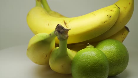Gelbe-Reife-Bananen-Und-Limetten-Rotieren-Auf-Dem-Tisch-Mit-Weißem-Hintergrund---Nahaufnahme